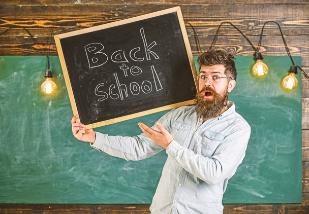 High school concept. Teacher in eyeglasses holds blackboard with title back to school. Man with beard and mustache on surprised face welcomes students, chalkboard on background - Фото, изображение