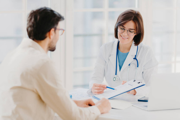 Medical appointment. Female doctor gives professional medical help to male patient, explains written information on paper in clipboard, gives support and good service, pose at hospital near desktop. - Photo, Image