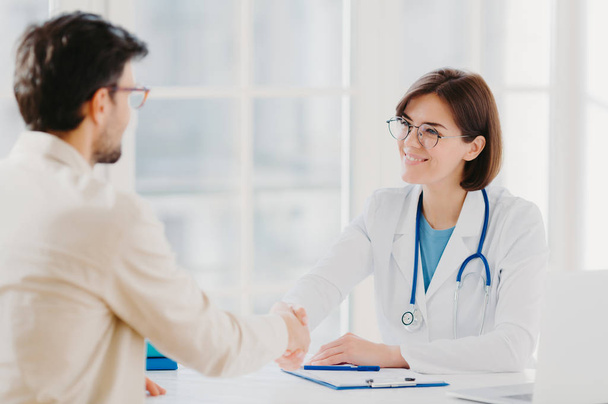Friendly female doctor greets patient with handshake, pose in private clinic, sit opposite each other, have talk, agree to sign insurance contract, meet in hospital. Health care and medicine concept - Photo, image