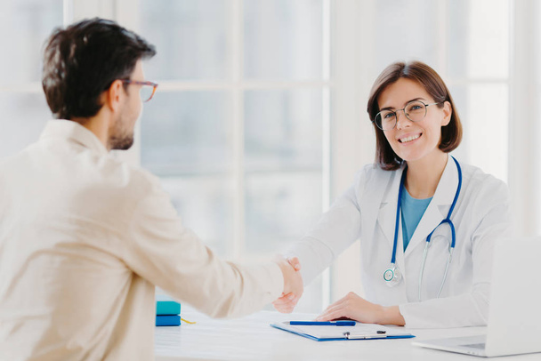 Partnership, assistance, trust and medicine concept. Female doctor shakes hands with thankful patient for good treatment and professionalism, pose in clinic, medical records near on white table - Foto, afbeelding