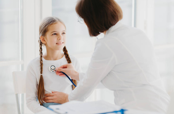 Caring female pediatrician listens childs lungs or heart with phonendoscope. Little girl comes on examination in private clinic, has every year doctor visit, explains symptoms of her disease - Foto, imagen