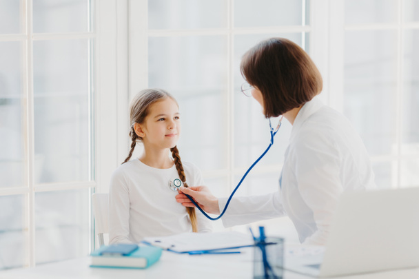 Woman pediatrician gives consultation to small girl, talk about health and symptoms, listens heart with phonendoscope, makes prescription, pose in hospital office. Childrean medical insurance and care - Fotografie, Obrázek