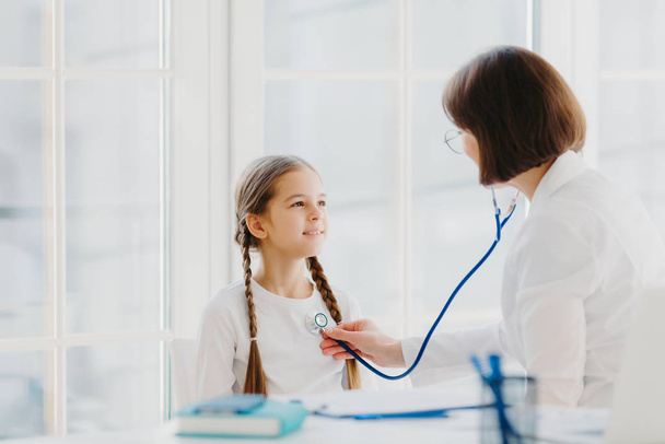 Lovely small girl listens consult of professional experienced doctor who listens her lungs with stethoscope, comes on medical checkup appointment. Children healthcare and clinic visit concept - Photo, Image