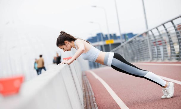 Young sports woman in fitness suit is pushing up from curb, parapet of bridge, against the backdrop of urban cityscape. - Fotografie, Obrázek