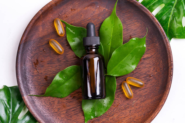 Naturalne produkty kosmetyczne z kapsułkami żelowymi Omega-3 i serum w szklanych butelkach, zielone liście na drewnianym talerzu na białym tle. - Zdjęcie, obraz