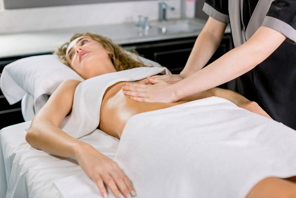 Side angle view of hands massaging female abdomen. Therapist applying pressure on belly. Pretty young blond woman receiving manual massage at spa salon - Photo, image