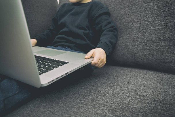 The child is sitting on the sofa holding the laptop and watches  - Foto, afbeelding