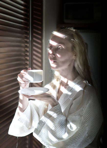 woman in a bathrobe with a cup of coffee by the blinds window in striped light - Foto, Bild