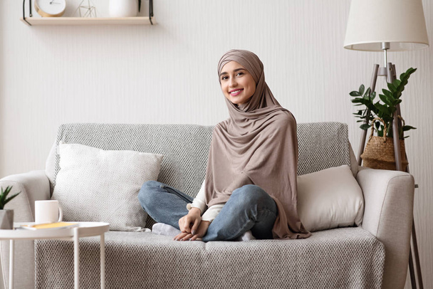 Ευτυχισμένο αραβικό κορίτσι με μαντίλα που ποζάρει στον καναπέ στο σπίτι - Φωτογραφία, εικόνα