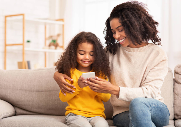 African Mother And Daughter Using Smartphone Sitting On Couch Indoor - Фото, зображення