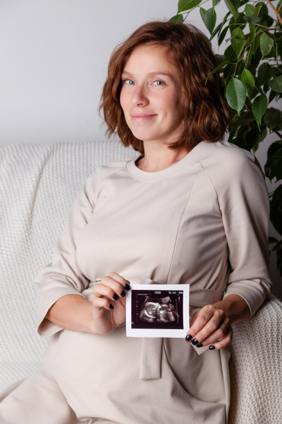Pregnant woman holding ultrasound image. Concept of pregnancy, health care, gynecology, medicine. Young mother waiting of the baby. Close-up, copy space, indoors. - Photo, Image