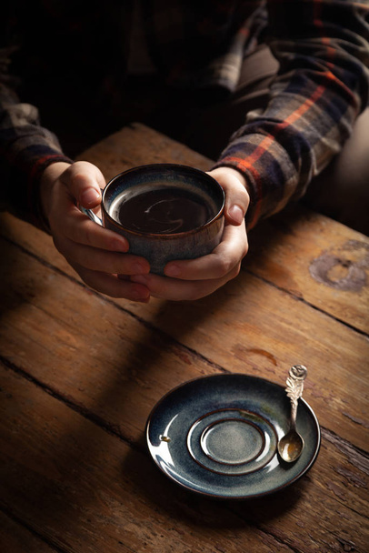 Крупный план черный американский кофе в синей чашке в мужской руке, блюдце со старинной чайной ложкой на деревянном коричневом винтажном фоне. Концепция завтрака в кофейне
 - Фото, изображение
