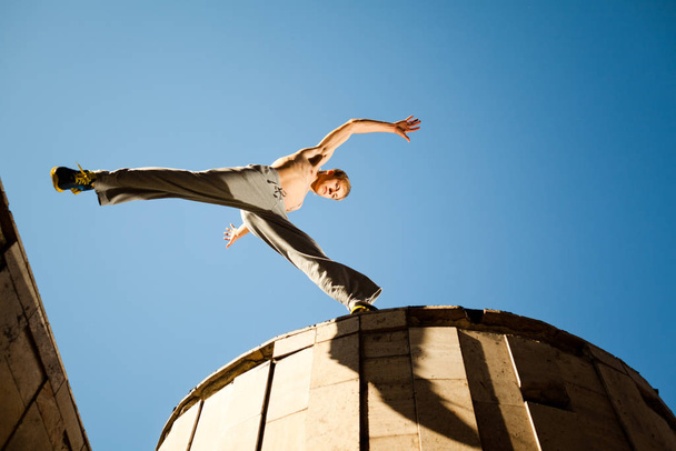 Young man jumping and practicing parkour between buildings roofs outside on clear summer day with blue sky at background - Foto, Imagen