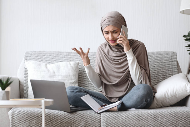 Evde dizüstü bilgisayar kullanırken cep telefonuyla konuşan endişeli müslüman kız. - Fotoğraf, Görsel