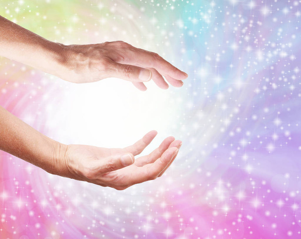 Creating a Healing Rainbow Energy Vortex from White Light - female cupped hands with a spiralling rainbow coloured sparkling background and a ball of white light plus copy space  - 写真・画像