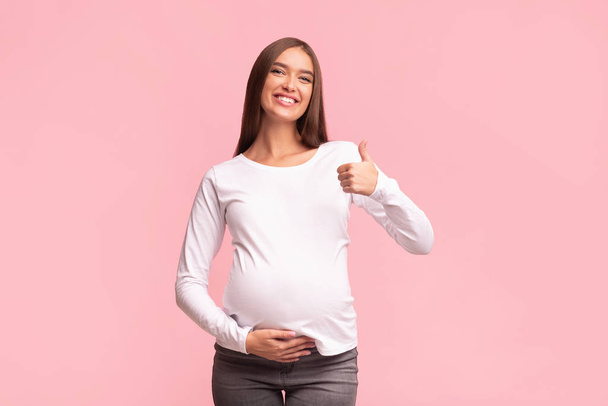 Vrolijke Zwangere Vrouw Gesturing Duimen-Up Poseren over roze achtergrond - Foto, afbeelding