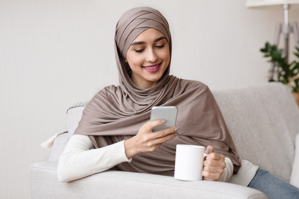 Smiling Girl In Hijab Resting At Home With Smartphone And Coffee - Фото, изображение