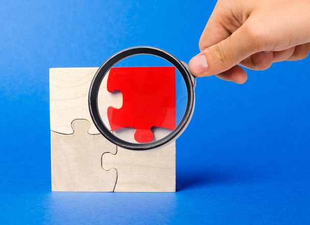 A magnifying glass looks at a red wooden puzzles on a blue background. Individual opinion. Stand out from the crowd. Uniqueness. Divergent views. Different concepts to other people. - 写真・画像