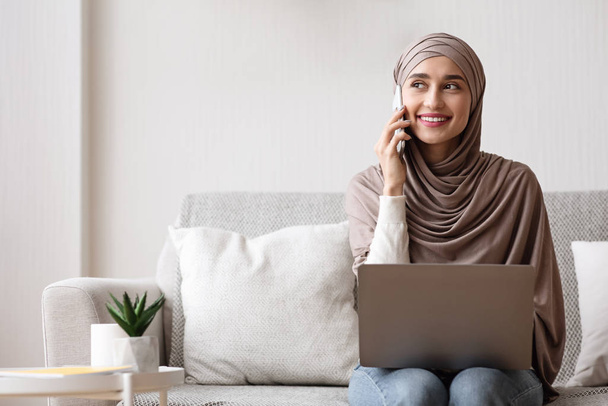 Evde dizüstü bilgisayar kullanırken gülümseyen Müslüman kadın cep telefonuyla konuşuyor. - Fotoğraf, Görsel