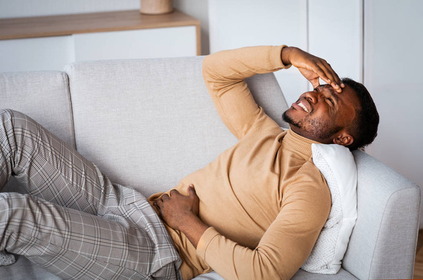 Uomo toccando Aching stomaco e testa sdraiato sul divano al coperto
 - Foto, immagini