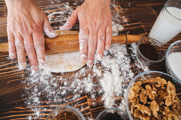 Woman is rolling pin on dough in kitchen, dough recipe ingredients and rolling pin on vintage wood table from above. - Фото, изображение