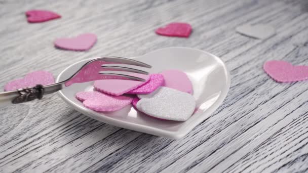 Dessert fork stirs a bunch of pink and white decorative valentines in a white plate in the shape of a heart on a light textural wooden table. Happy Valentine's Day Greetings - Кадры, видео