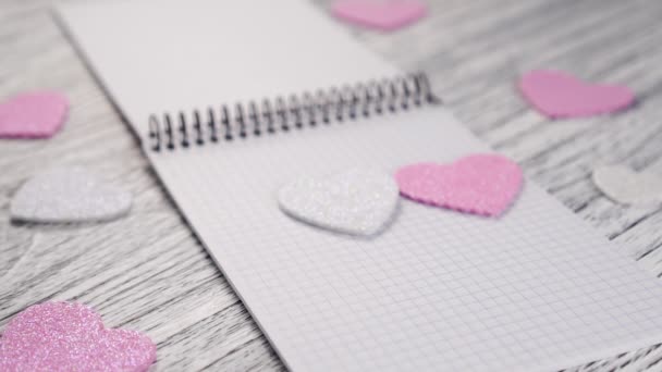 Błyszczące papierowe serca spadają na otwarty notatnik z czarnym metalowym wiązaniem na białym drewnianym stole teksturowym. Pomysł gratulacji w Walentynki i deklaracja miłości - Materiał filmowy, wideo