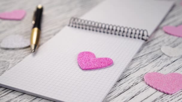 Блискучі рожеві та білі паперові серця падають на відкритий ноутбук з чорним металевим зв'язуванням. З чорною жовтою ручкою на білому дерев'яному текстуровому столі. Ідея привітання у Валентинів день і декларація любові - Кадри, відео