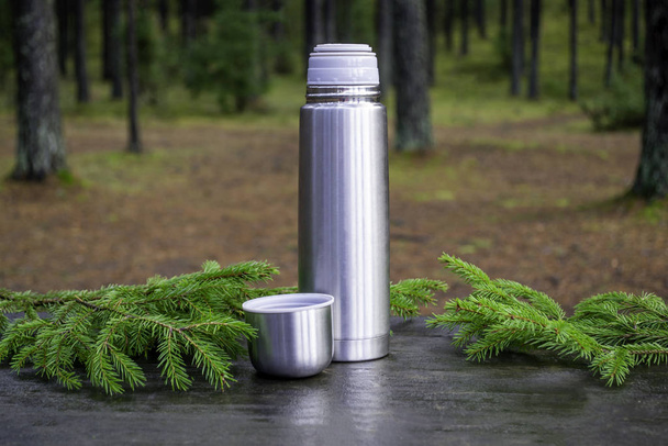 Thermos with an open lid that stands next to it. on a wooden surface with fir branches. Nature on background. The concept of outdoor recreation in the cold season. - Photo, Image