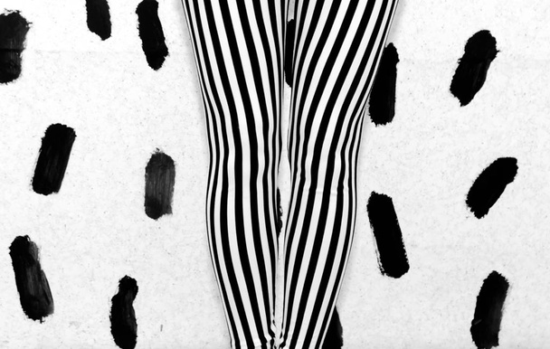 De benen van het meisje in zwart-wit gestreepte leggings, parallel aan elkaar gelegen tegen een witte muur bedekt met zwarte penseelstreken - Foto, afbeelding