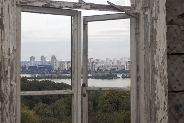 Θέα από ένα παλιό σπασμένο παράθυρο στην τοπογραφία του πάρκου, ένα ποτάμι με νησιά και πολυώροφα σύγχρονα κτίρια στην απέναντι όχθη - Φωτογραφία, εικόνα