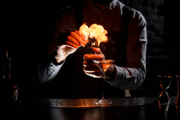 Bartender spraying on a lighter with orage zest juice to golden alcoholic drink in a martini glass - Fotografie, Obrázek