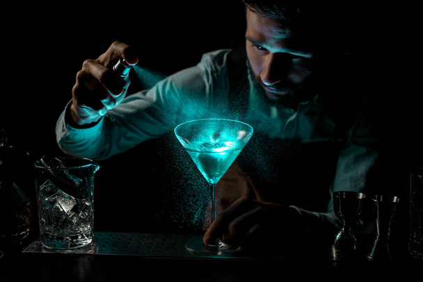 Μπάρμαν ψεκασμό με ένα πικρό σε ένα μπλε αλκοολούχο κοκτέιλ σε ένα ποτήρι μαρτίνι διακοσμημένα με ένα spikelet - Φωτογραφία, εικόνα