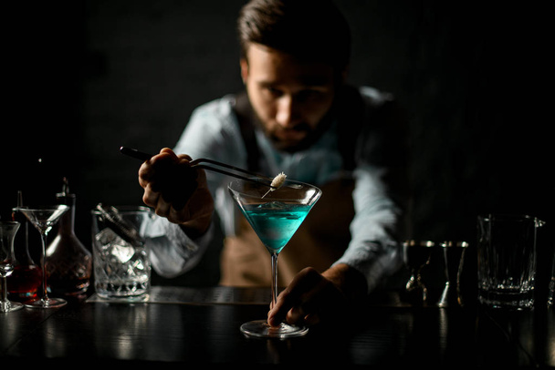 Professional bartender deacorating a blue alcoholic cocktail in a martini glass with a spikelet with twezzers - Foto, Imagem