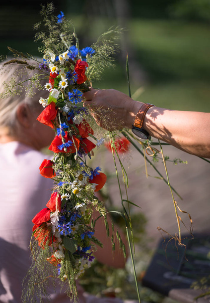 duas mãos de mulheres tecem a grinalda, tradição de solstício de verão, grinalda de flores de campo, grama e cereais, processo de tecelagem de grinalda
 - Foto, Imagem