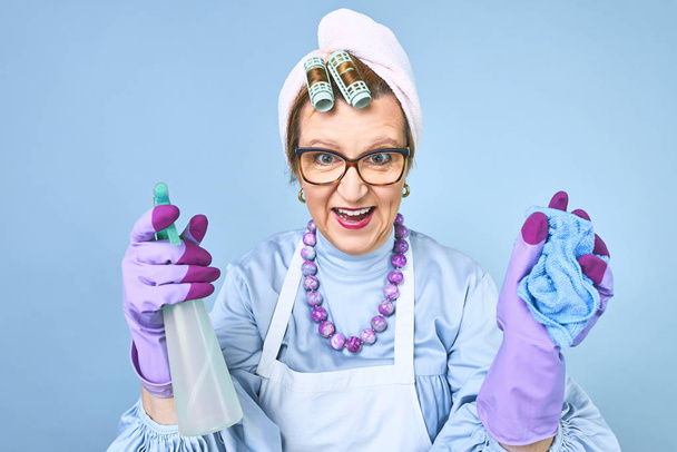 Portrait of happy old cleaning woman in apron with dust cleaning brush isolated on blue background. special uniform and professional equipment for cleaning - Photo, Image