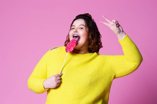 Cheerful plus size model with big lollipop, fat woman in yellow jumper on pink background - Фото, зображення