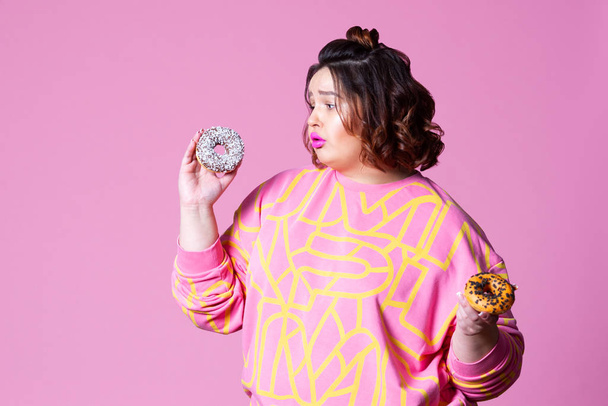 Sad plus size model with doughnuts, fat woman sadness and overeats on pink background - Foto, imagen