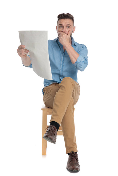 wstrząśnięty młody człowiek czytający gazetę i zakrywający usta  - Zdjęcie, obraz