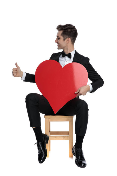 smiling elegant groom holding big red heart and making ok sign - Fotó, kép