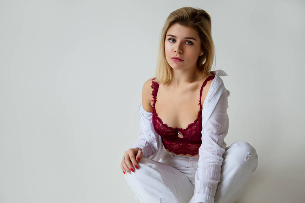 Beautiful blonde girl posing in studio in lace red lingerie, white jeans and white shirt, sexy, white background, daylight - Φωτογραφία, εικόνα