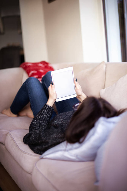 Rear view shot of woman lying on sofa at home and holding digital tablet wiht blank screen in her hands. - Foto, immagini
