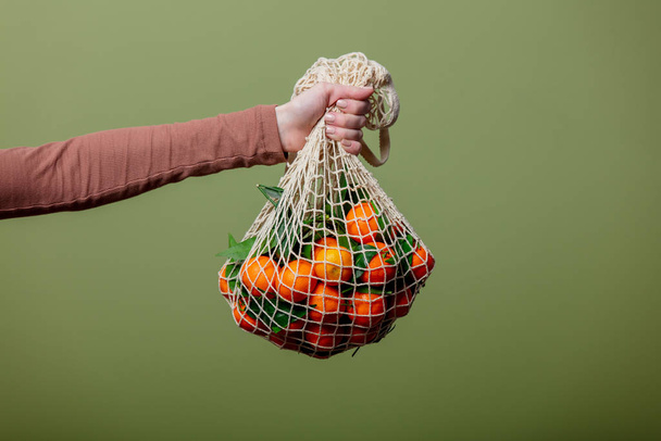 Woman hand holds mandarins in a string bag on green background - Fotó, kép