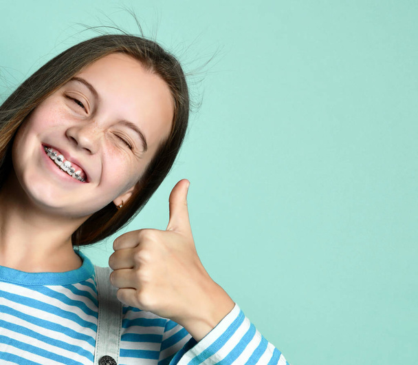 Teenage female in jeans overall, striped sweatshirt. She smiling, showing thumbs up, winking, posing on blue background. Close up - Fotó, kép