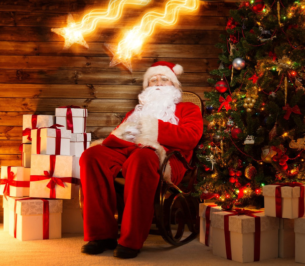Père Noël assis sur une chaise à bascule à l'intérieur de la maison en bois avec des boîtes-cadeaux autour de lui et deux étoiles tombantes
 - Photo, image