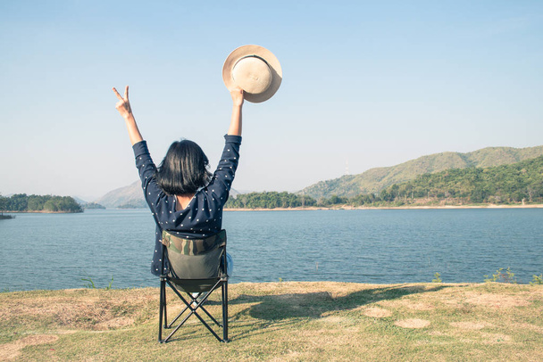 Urlaubs- und Reisekonzept: Asiatin trägt Hut und sitzt entspannt auf tragbarem Stuhl am Nationalpark. - Foto, Bild