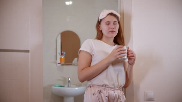 Μια νεαρή γυναίκα με πιτζάμες πίνει καφέ στην πόρτα του μπάνιου το πρωί. - Πλάνα, βίντεο