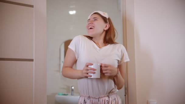 Egy fiatal, mosolygó nő táncol reggel a fürdőszoba ajtajában egy csésze kávéval a kezében. - Felvétel, videó