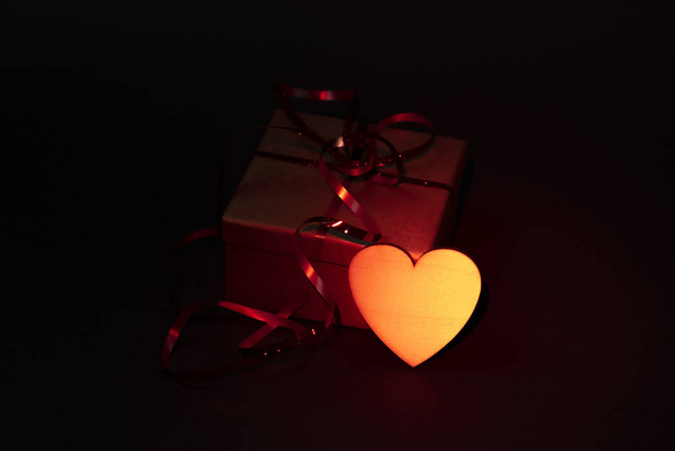 Caja de regalo con una cinta roja y un corazón dorado sobre un fondo gris. Retroiluminación roja
 - Foto, imagen
