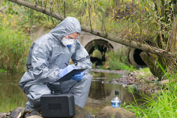 a specialist, in a protective suit and gloves, working with toxic substances, records the results of a sample of liquid from the river, on a sheet on a tablet, against the background of cleaning pipes - Photo, Image